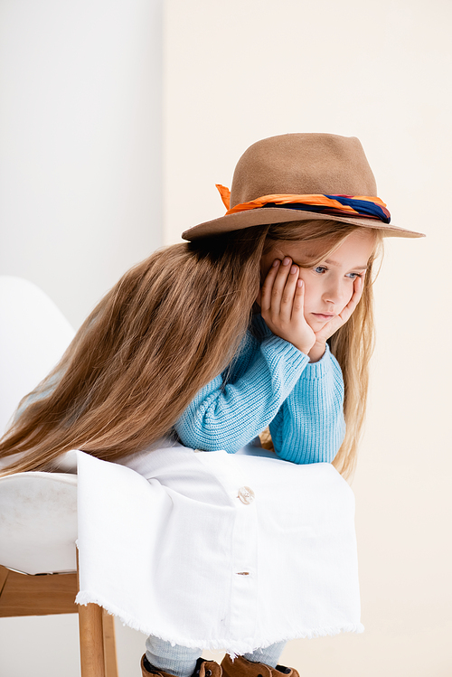 sad fashionable blonde girl in brown hat, white skirt and blue sweater sitting on chair near beige wall