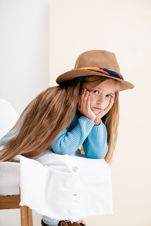sad fashionable blonde girl in brown hat, white skirt and blue sweater sitting on chair near beige wall