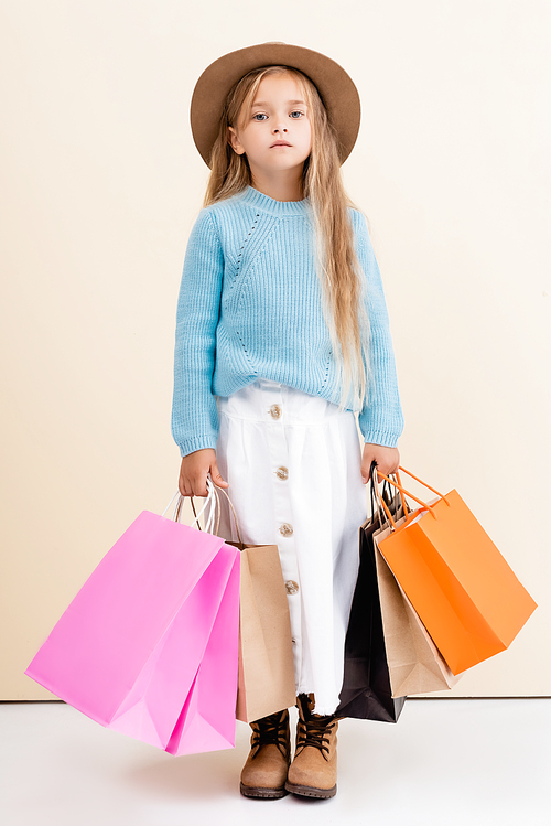 fashionable blonde girl in brown hat and boots, white skirt and blue sweater with colorful shopping bags near beige wall