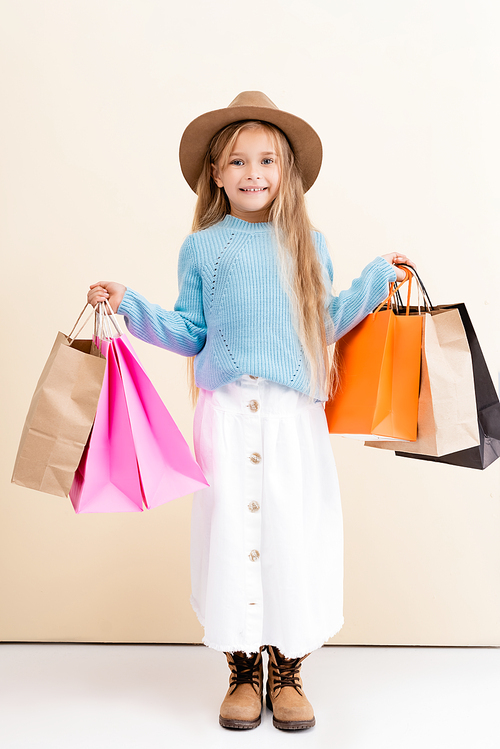 fashionable blonde girl in brown hat and boots, white skirt and blue sweater with colorful shopping bags smiling near beige wall