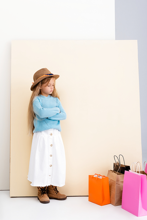 sad fashionable blonde girl in brown hat and boots, white skirt and blue sweater near colorful shopping bags and beige wall