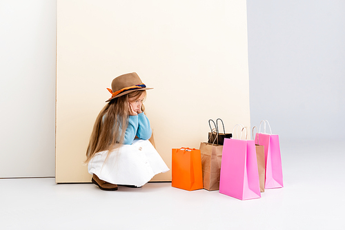 sad fashionable blonde girl in brown hat and boots, white skirt and blue sweater sitting near colorful shopping bags and beige wall