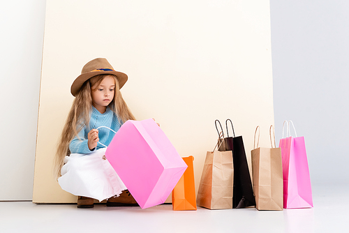 fashionable blonde girl in brown hat and boots, white skirt and blue sweater looking inside colorful shopping bag near beige wall