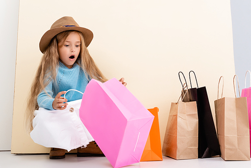 shocked fashionable blonde girl in brown hat and boots, white skirt and blue sweater looking inside colorful shopping bag near beige wall