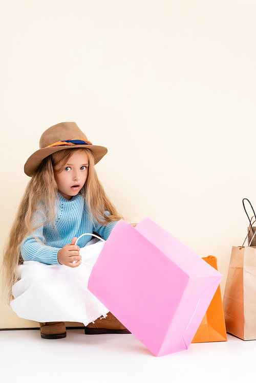 shocked fashionable blonde girl in brown hat and boots, white skirt and blue sweater looking inside colorful shopping bag near beige wall