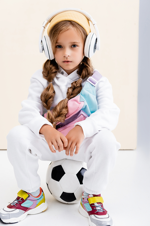 blonde girl in sportswear and headphones posing with soccer ball on beige and white background