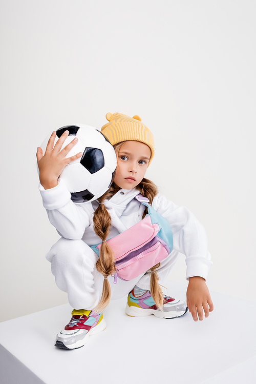 blonde girl in sportswear posing with soccer ball on cube isolated on white