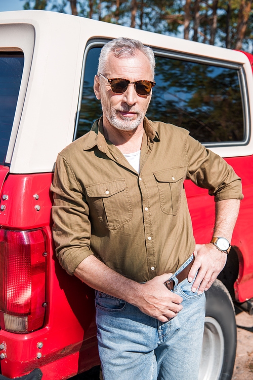 confident senior man in sunglasses standing with hand on hip near red car and 