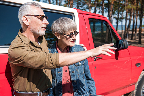 senior couple in sunglasses standing near red car in sunny day