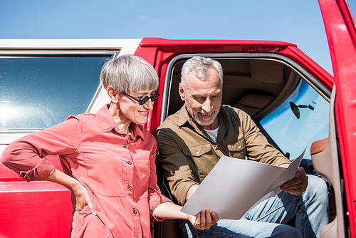 smiling senior traveler sitting in red car and looking at map with wife