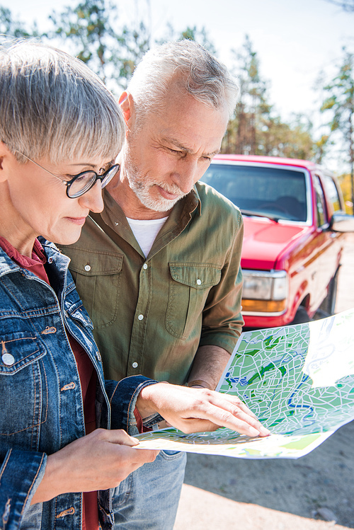 senior couple of tourists standing near car and looking at map in sunny day