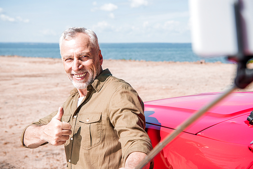 smiling senior man standing near red car and taking selfie while showing thumb up in sunny day