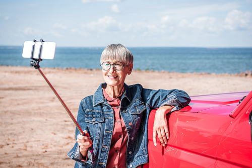 smiling senior woman in denim jacket standing near red car and taking selfie in sunny day