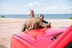 back view of senior couple standing near red car at beach and embracing in sunny day