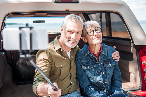 smiling senior couple sitting on car, embracing and taking selfie