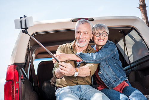 smiling senior couple sitting on car, embracing and taking selfie