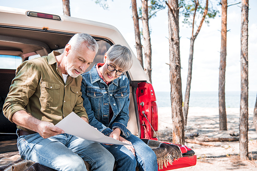 smiling senior couple sitting on car and holding map in sunny day