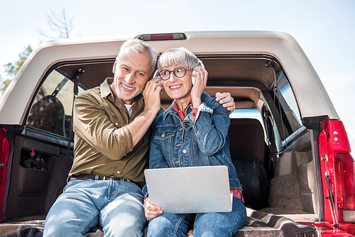 smiling senior couple of tourists with laptop listening music in headphones in car