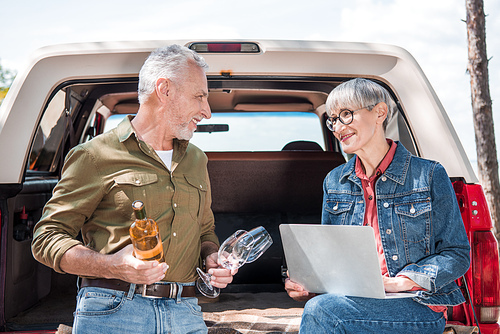senior man holding wine and wine glasses and looking at wife using laptop near car