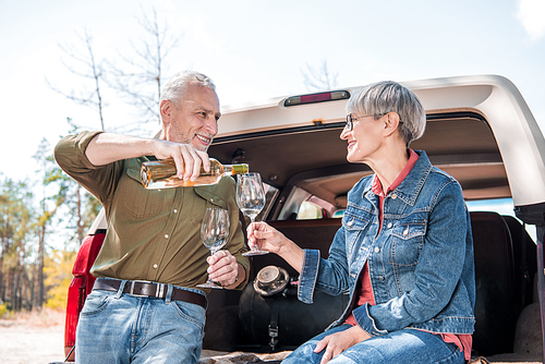 smiling senior man pouring wine in wine glasses and looking at wife near car