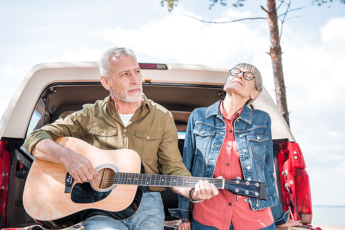 senior man playing acoustic guitar while standing near car with wife
