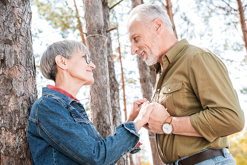 happy senior couple holding hands and looking at each other near car in forest