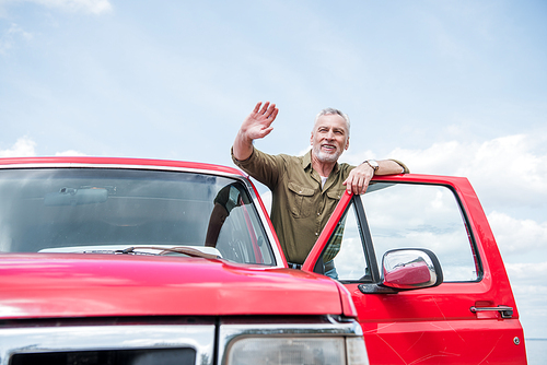 senior man in shirt in red car waving hand and smiling in sunny day