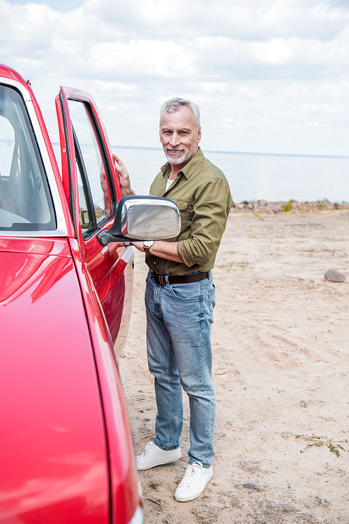 full length view of smiling senior man standing near red car at beach