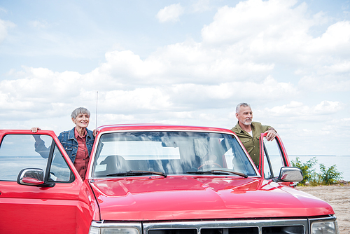 senior couple of tourists standing near red car at beach