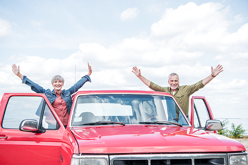 smiling senior couple of tourists standing with hands up near red car at beach