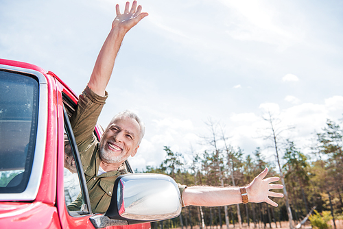 excited senior man in red car waving hands under blue sky