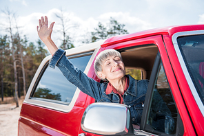 happy senior woman waving hand from car window with closed eyes