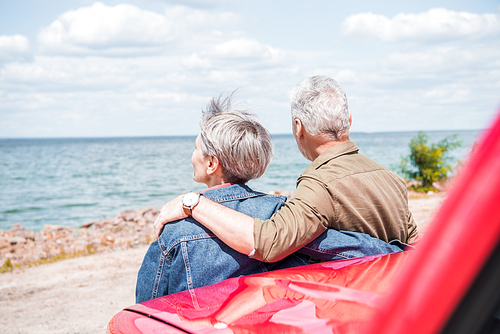 back view of senior couple standing near red car and embracing at beach