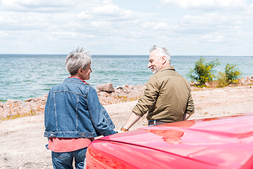back view of senior couple standing near red car and looking at each other at beach