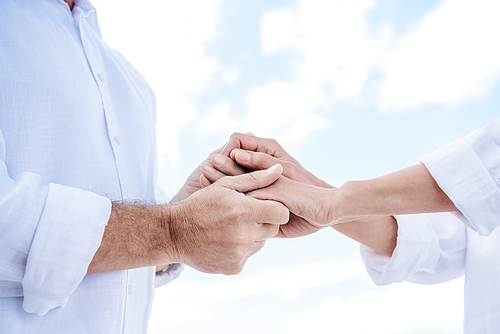 cropped view of senior couple in white shirts holding hands under blue sky