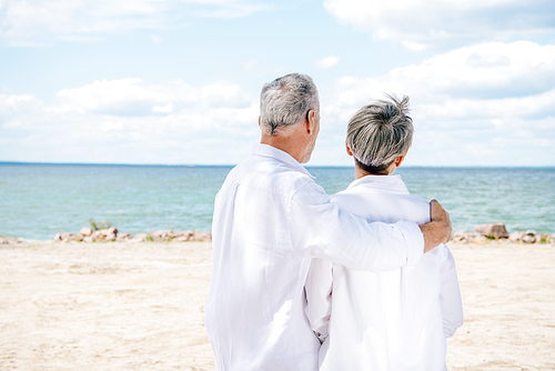 back view of senior couple in white shirts embracing at beach