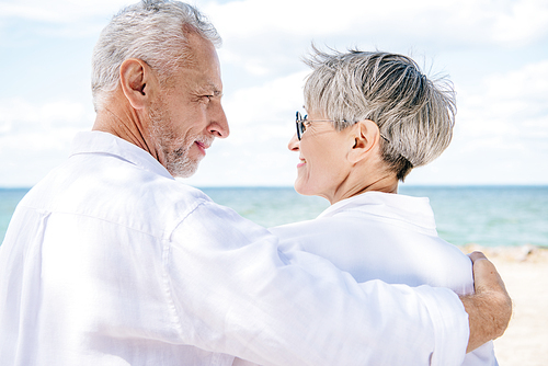 smiling senior couple in white shirts embracing and looking at each other near river
