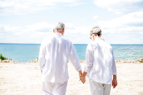 back view of senior couple in white shirts holding hands at beach