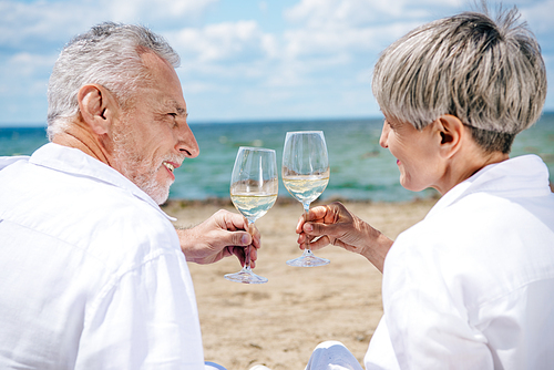 smiling senior couple holding wine glasses with wine and looking at each other at beach