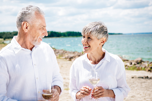 smiling senior couple holding wine glasses with wine and looking at each other at beach