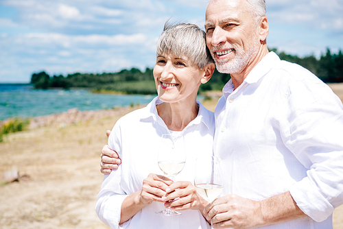 smiling senior couple embracing and holding wine glasses with wine at beach