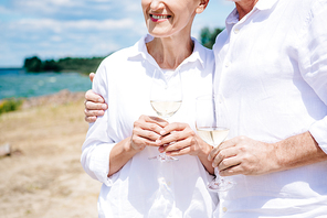 cropped view of smiling senior couple embracing and holding wine glasses with wine at beach