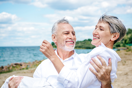 smiling senior man in white shirt lifting wife at beach under blue sky