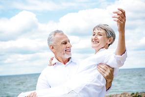 smiling senior man in white shirt holding wife under blue sky near river