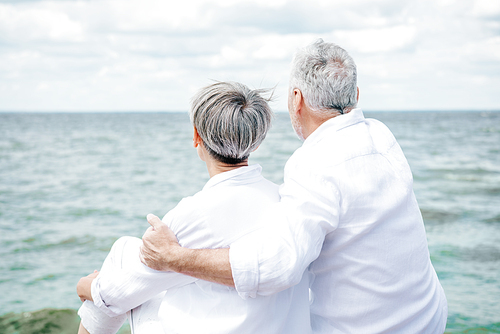 back view of senior couple in white shirts embracing near river under blue sky