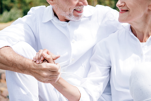 cropped view of smiling senior couple in white shirts holding hands