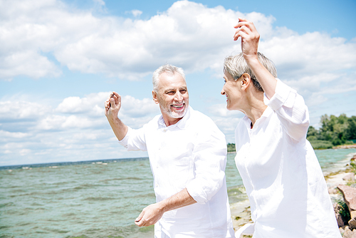 happy smiling senior couple in white shirts looking at each other and gesturing at beach under blue sky