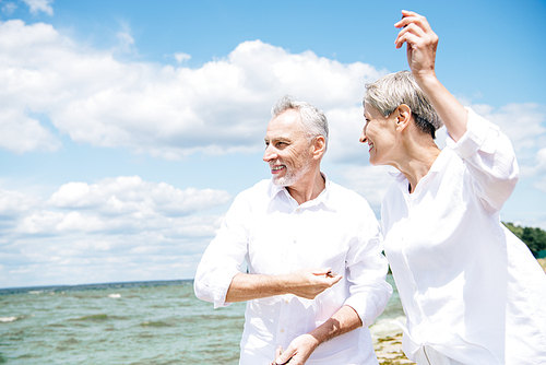 happy smiling senior couple in white shirts gesturing at beach under blue sky