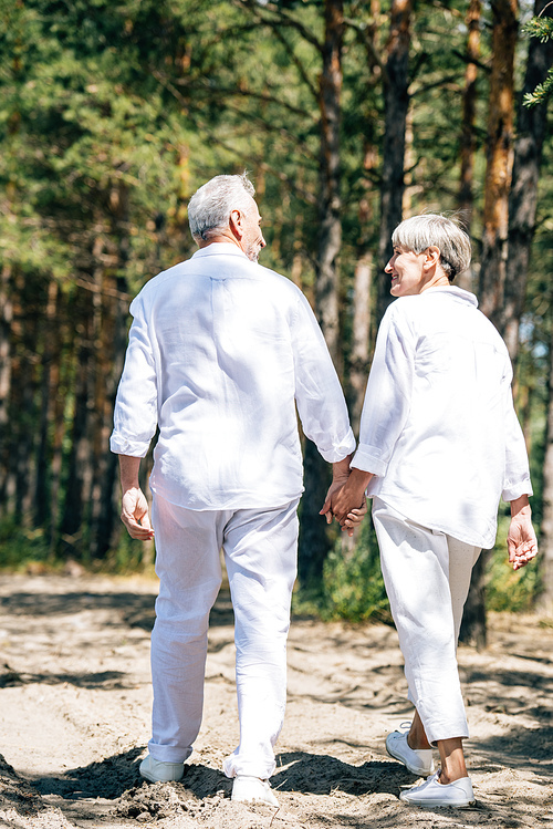 back view of happy senior couple in white shirts holding hands and looking at each other in forest