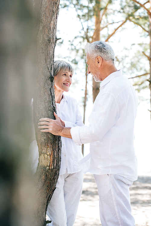 smiling senior couple standing near tree and looking at each other in forest